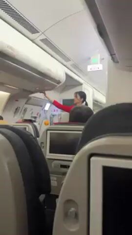 Here's a description of the video. 

The scene is inside an airplane cabin. The camera is positioned from a passenger seat, facing forward. An Asian woman, presumably a flight attendant, is standing in the aisle, reaching up to adjust something inside an overhead compartment. She's wearing a red uniform with black hair pulled back in a ponytail. Passengers are seated in the rows in front of and around the camera, visible by their headrests. Each seat is equipped with a personal video screen and tray. An "EXIT" sign is visible above the overhead compartment. The interior of the plane is clean and modern. The light is bright from the overhead lights.