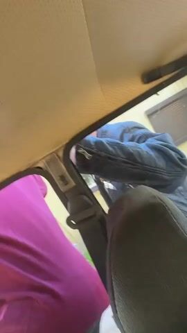 The video shows the interior of a car, likely from the driver's seat. A man in a blue jacket is seen outside the driver's side window, bending down as if interacting with someone inside. Another person, wearing a maroon shirt, is partially visible in the driver's seat. The focus shifts to the car's interior, revealing a close-up of a textured gray surface, possibly the seat or interior paneling. The environment outside the car seems to be a residential area with a yellow building visible.