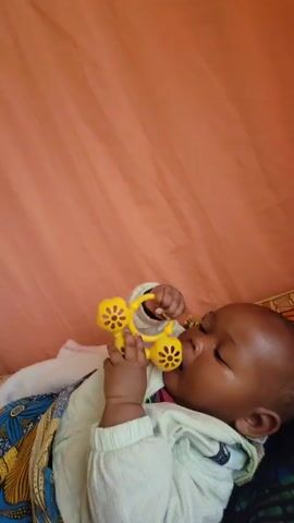 A baby lies on its back, partially wrapped in a colorful blanket and wearing a light green garment. The baby is holding a yellow plastic toy in its hand and has it near its mouth. The baby's eyes are mostly closed. The background is a plain peach-colored surface. The baby is making a vocalization, possibly crying.