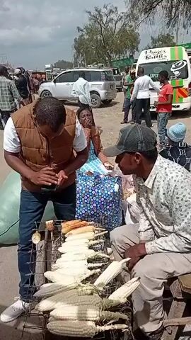 The video shows a man standing outdoors near a grill laden with corn on the cob. He's wearing a white t-shirt, a brown vest, blue jeans, and white sneakers. He appears to be interacting with his phone or some small object in his hands. Next to him are bags, one large and green, and another with a blue and white pattern. In the background, there's a bustling scene with people walking around, vehicles parked, and buildings in the distance, suggesting a public market or roadside vendor area.