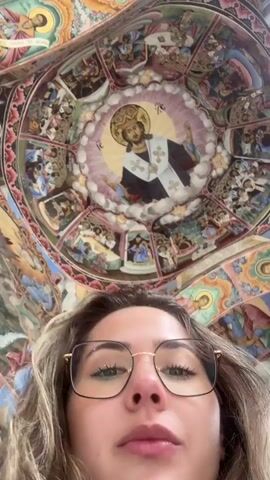 The video shows a person looking up at an ornate, colorful ceiling covered in religious paintings and images. The ceiling is circular in design with a central image of a religious figure surrounded by other scenes and portraits. The person has light brown, curly hair and is wearing gold-rimmed glasses. Their face is visible in the foreground, looking upwards towards the ceiling. The lighting is bright, illuminating the ceiling's details. The camera is held at a low angle, emphasizing the height and intricacy of the ceiling.