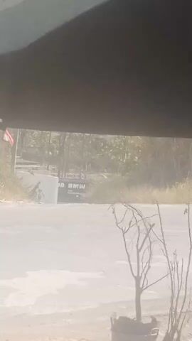 The video shows a view from under a dark overhang, looking out at a hazy street scene. On the left, a white concrete barrier is visible, with what appears to be a sign in the background with the text "GOOD BMW." The road is paved and appears wet in places. To the right of the road, there is vegetation and a bare-branched tree in a pot in the foreground. The overall impression is of an outdoor scene on a bright, possibly dusty or humid day.
