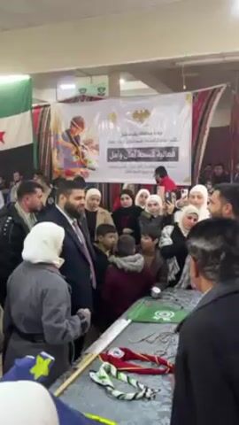 The video shows a crowded indoor event with a backdrop featuring a poster with Arabic script and a drawing of a young boy with a tool in his hand. There are flags on either side of the poster. A large group of adults and children are gathered around a table. Many are wearing headscarves, and a few men are in suits. People are taking photos with their phones. A woman in a gray coat points towards something on the table. Several items are visible on the table, including green flags and a neckerchief. The overall atmosphere appears to be celebratory or ceremonial.