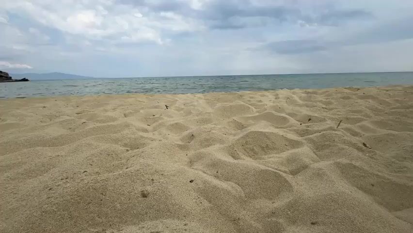 A low-angle, static shot of a sandy beach in front of a calm sea and a cloudy sky. The sand in the foreground is rippled and textured. The sea is a dark blue-green with gentle waves. The sky is filled with scattered clouds, some of which are white and fluffy, while others are darker and more ominous. In the distance, there is a faint outline of land and a rocky outcrop on the left side of the frame. There are no people or objects visible in the scene.