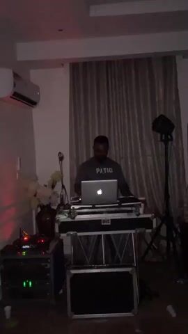 The video shows a man standing behind a DJ setup in a dimly lit room. He is wearing a gray t-shirt with the word "PATIO" printed on it. In front of him is a laptop, a DJ controller, and other audio equipment. To his left, there is a vase with flowers and a rack of electronic equipment with green lights. To his right, there is a speaker on a stand. The room has beige curtains in the background and an air conditioner on the wall. The man appears to be operating the DJ equipment, with his hands on the controller.