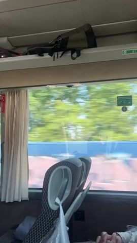 The video shows the interior of a bus or train. The camera is positioned behind a row of seats, looking out a window. Outside the window, trees and buildings are visible, and the scenery is blurred, indicating movement. Overhead compartments hold bags and other items. A red fire extinguisher is mounted on the wall near the window.