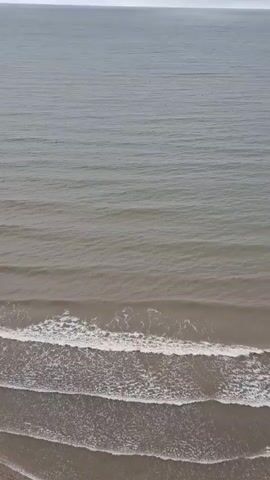 This media file is a drone shot video overlooking a beach and the Baltic Sea in Sääre, Estonia. The scene is dominated by the vast expanse of the ocean, which appears calm with gentle waves rolling towards the shore. The water is a muted grey-blue, reflecting the overcast sky. The beach is visible in the foreground, a strip of sand with a few faint ripples left by the receding tide. The horizon is a hazy line where the sea meets the sky.

The activity taking place is the natural movement of the sea, with waves consistently breaking on the shore. There are no visible people or man-made objects in the frame, emphasizing the natural beauty and solitude of the location. The weather appears to be cloudy, contributing to the soft, diffused light and the muted color palette of the scene. The time of day is indeterminate due to the overcast conditions, but it seems to be daytime. The overall mood is one of tranquility and peacefulness, showcasing the serene coastal landscape of Estonia. There is no visible text in the media file.