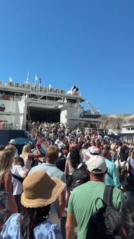 The video shows a large crowd of people boarding a ferry. The ferry is white with a blue stripe near the waterline. The ramp to board the ferry is open, and people are walking up it. Many of the people are carrying bags. The weather appears to be sunny and clear, with a bright blue sky. The ferry is docked at a port, with rocky hills visible in the background. There is another boat docked next to the ferry, also filled with people. The scene suggests a busy travel day.