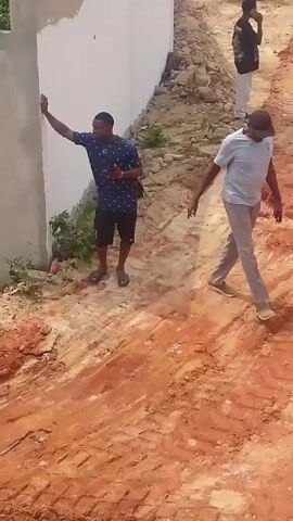 The video shows a construction site with three men. One man is standing near a white wall, appearing to take pictures. Another man is in the background, holding something to his ear. The main subject is a man in a gray shirt and cap, walking across a reddish-brown dirt area that shows tire tracks. The camera's viewpoint is from above and slightly behind a concrete block or structure. The sound of heavy machinery is present.