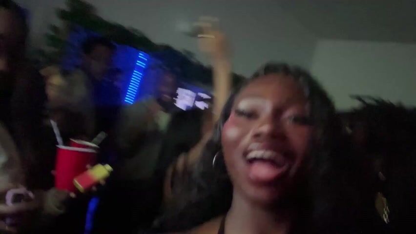 Three young women are taking a selfie. Two of them are in focus, while the third is partially obscured in the background. The woman in the front left is of Asian descent and has long dark hair. She is wearing a leopard-print top. The woman on the front right is Black and has long dark hair and is wearing a black top. Both women are smiling and appear to be enjoying themselves. The background is blurry and indistinct but appears to be a party setting. Blue lights are visible in the background, adding to the festive atmosphere.