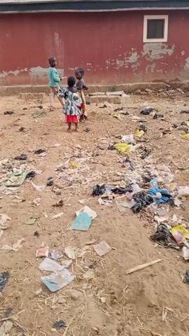 The video shows an open area with sandy ground heavily littered with garbage. A red building with a window is visible in the background. Three young children are present: two girls in patterned clothing and a boy in a blue shirt and beige pants. They stand near the building, seemingly interacting playfully.  A man pushes a wheelbarrow along a dirt road towards the background. Palm trees are visible further down the road.