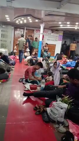The video depicts a large indoor space, possibly a waiting area or temporary shelter, filled with people. Most are lying down on mats or blankets, many seemingly resting or sleeping. The room appears crowded, with belongings like bags and water bottles scattered around.  The lighting is somewhat dim and there are several ceiling lights visible. People are wearing a variety of clothing, including traditional garments. Some individuals are sitting, talking, or attending to personal belongings. A person wearing a pink shirt walks across the foreground. The overall atmosphere appears to be one of temporary accommodation or waiting.