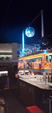 The video shows the interior of a dimly lit bar or nightclub. In the first few seconds, a disco ball is visible hanging from the ceiling, reflecting light. Shelves behind a bar are stocked with various bottles of alcohol and illuminated from below. In the background, people are seen moving around and interacting. As the video progresses, the lighting changes, with blue and purple lights becoming more prominent. There are shots of people at the bar, some with drinks in hand. The overall atmosphere appears to be lively and social.