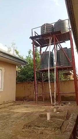 The video shows a residential scene with a water tower in the background. The water tower is constructed of red painted steel and has two levels, each supporting two large black water tanks. A white ladder is affixed to the structure. Adjacent to the water tower is a single-story building with a tan exterior and a visible window. The ground is mostly concrete, with some scattered dirt and debris. A low wall borders the area. Above, the sky is blue with scattered clouds. A tall tree is visible between the building and the water tower, partially obscuring the view. The camera pans slightly upward and to the right, revealing the building's roof and a tree.