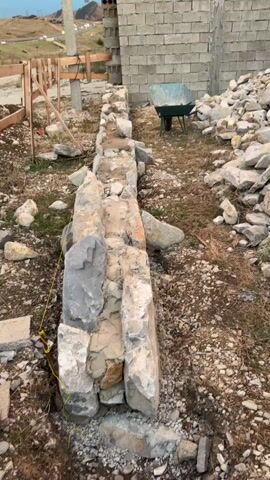 The video shows an outdoor scene during the day. In the foreground, there is a low wall constructed of large, irregular stones. Some of the stones are rough and unfinished, while others appear to be more shaped. The wall is partially filled with mortar. To the right of the wall, there is a large pile of loose stones and rubble. Further back, a partially constructed building is visible, made of concrete blocks. A green wheelbarrow is positioned near the building. In the middle ground, a cat with tabby markings and white paws is walking across the rough terrain, which is covered with dirt, small stones, and dry grass. The background shows a hillside with some vegetation and distant structures. The overall impression is a construction site or an area undergoing renovation.
