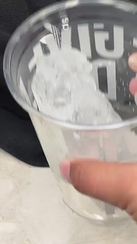 A clear plastic cup containing a large piece of ice sits atop a white surface next to a pair of black Adidas sweatpants. A hand is briefly visible next to the cup.