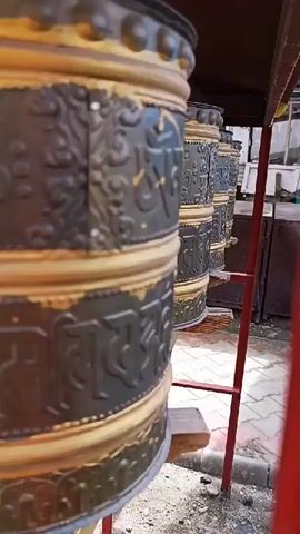 A shallow depth of field shot shows a row of Tibetan prayer wheels, the closest one in focus and slightly left of center. The prayer wheels are cylindrical and appear to be made of metal with ornate designs and script. They are arranged in a line, with some of them partially obscured by the ones in front. The prayer wheels are supported by wooden structures and are mounted beneath a wooden overhang. A red metal ladder is visible to the right of the prayer wheels. The ground beneath the prayer wheels is a mixture of gravel and paving stones. The overall scene suggests an outdoor location, possibly a temple or monastery.