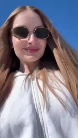 The video features a woman, likely in her late 20s to mid-30s, with long, straight light brown hair, wearing sunglasses and a white hoodie. She is standing outside on what appears to be a paved road. In the background, there is a snow-covered landscape with a few houses and trees visible. The sky is clear and blue. The camera is held close to the woman's face, suggesting she is filming a selfie video. She is smiling slightly and appears to be moving, as the background subtly shifts. The overall setting seems to be a rural or semi-rural area in early spring, where snow is still present.