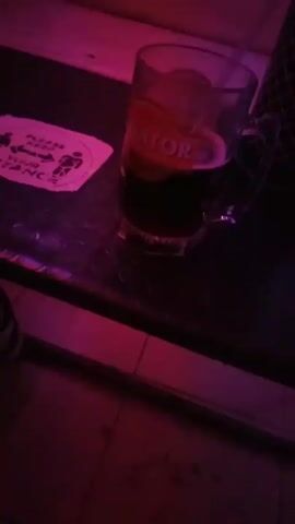 Here's a detailed description of the video:

**Setting and Environment:**
The video appears to be filmed inside a dimly lit bar or lounge, illuminated predominantly by pink/purple neon lights. The flooring is tiled.

**Objects:**
*   A glass mug filled with a dark liquid (possibly a beer or cocktail) sits on a table. The mug has the word "Senator" printed on it. A straw can be seen in the glass.
*   A white coaster with a social distancing message is also on the table next to the mug.
*   There's a water bottle with a black lid and a red container next to it on a light-colored cushioned bench.
*   There are lounge-style benches with white cushions.
*   Dark, small tables are arranged near the seating.
*   A TV screen is visible in the background, along with wall decorations.

**People:**
*   Several people can be seen in the background, including bar staff and customers.

**Actions and Interactions:**
*   The camera is moving, starting with a close-up of the drink and then panning around the room to show the seating area.
*   Background music is playing.
*   People in the background seem to be interacting with each other.

**Overall Impression:**
The video gives the impression of a casual, dimly lit bar or lounge atmosphere, with people relaxing and enjoying drinks. The color scheme is dominated by pink and purple lighting.