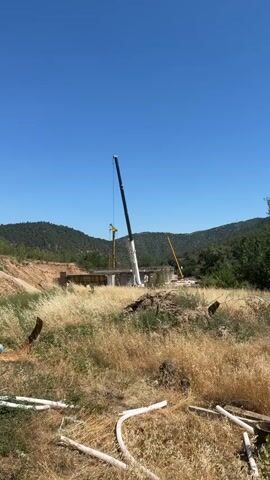 This is a vertical video with a bright blue sky and lush green hills in the background. In the foreground, dry, tall grass and some white plastic pipes are visible. In the middle ground, there is a construction site with a partially built bridge. A large white crane with a black boom is prominently positioned on the left side of the frame, with its boom extended over the bridge. To the left of this crane, a smaller yellow crane is also visible. Further to the right, another yellow crane with a long, slender boom stands tall. The construction site appears to be in a rural or mountainous area, with trees and vegetation surrounding it. The overall impression is one of ongoing construction work in a natural landscape.