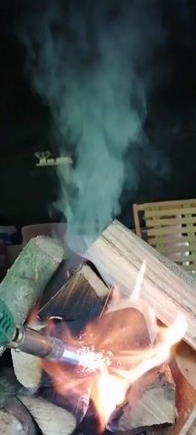 A person uses a handheld blowtorch to ignite a pile of firewood. The flames are orange and yellow, and the wood is placed on a metal grate inside a fireplace. Smoke rises from the burning wood. The blowtorch is green and attached to a red gas canister. A wooden crate is visible in the background.
