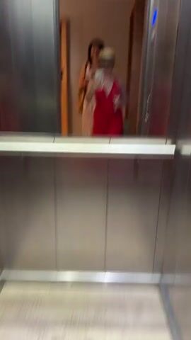 The video shows a group of people inside an elevator, reflected in a mirror. The primary subject is a woman in the foreground, wearing a red dress, a straw hat, and sunglasses. She is holding a smartphone, taking a selfie. She also appears to be holding a glass. Behind her, another woman is visible, wearing a white dress with a floral pattern, and holding a similar glass. A third person, possibly a man, is partially visible behind the woman in the red dress.

The activity taking place is the group entering or riding in the elevator, with the woman in front documenting the moment with a selfie. The setting is clearly an elevator, indicated by the metallic walls and the presence of elevator doors and buttons visible in the reflection.

Notable details include the women's attire, suggesting a casual or possibly celebratory occasion, perhaps a party or a night out. The bright red dress and the straw hat add a touch of style. The reflections in the mirror are slightly blurred, but the overall scene is clear. The lighting appears to be artificial, typical of an indoor setting like an elevator. There is no visible text in the video. The presence of drinks suggests a social gathering. The location context of Dračevice, Bosnia and Herzegovina, provides a geographical setting for this moment.