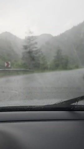 The video shows a view from inside a car looking through the windshield. It is raining heavily and the windshield wipers are intermittently clearing the view. The road ahead is wet and winding through a mountainous, forested area. The trees are green and appear to be pine or fir. The sky is overcast and gray. The overall impression is of a drive through a scenic but wet landscape.