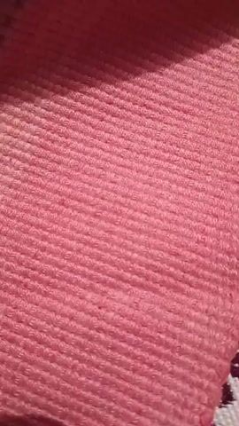 The video shows a close-up view of a textured pink fabric. The fabric has a ribbed pattern running horizontally across the screen. In the bottom right corner of the video, a small portion of a patterned fabric with dark purple and white geometric shapes is visible. The lighting in the video creates shadows that highlight the texture of the pink fabric. The camera appears to be held steady, with no noticeable movement.