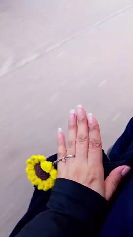 The video shows a person's hand, with neatly manicured fingernails, wearing a silver ring connected to a small, crocheted sunflower. The person is wearing a black long-sleeved shirt and blue pants. The background is a blurred, light grey surface that appears to be the ground, suggesting the person is moving. The video is shot from a first-person perspective, looking down at the person's own hand.