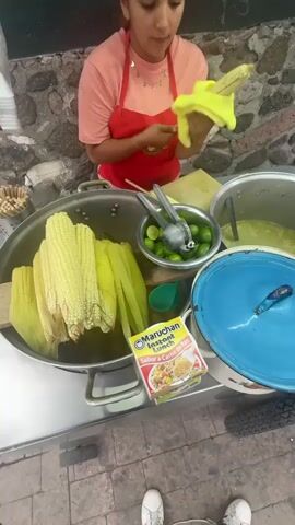 A young woman in a red apron is preparing corn on the cob from a food cart. The corn is in a large metal pot, still with the husk on. She is using a yellow towel to hold the corn while inserting a wooden skewer. There is a bowl of limes with a squeezer, a container with a greenish liquid and a blue-lidded pot.  A packet of “Maruchan Instant Lunch” in Spanish is visible on the cart. The cart is on a brick walkway in front of a stone wall. The camera is positioned to see the vendor and the food items on the cart.