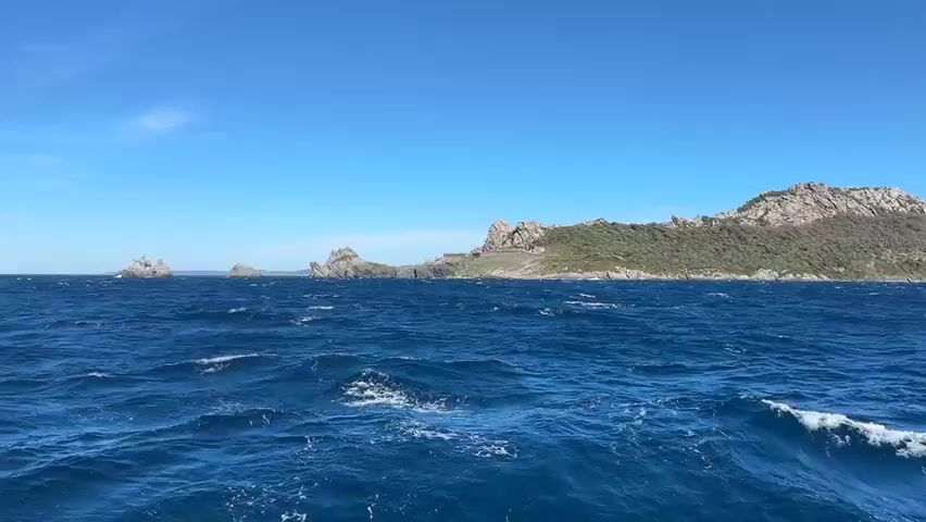 The video shows a view from a boat on the ocean. The water is a deep blue with whitecaps. In the distance, there is a rocky island with a fortress built on top of it. The sky is clear and blue. The camera is moving slightly, as if on a boat.