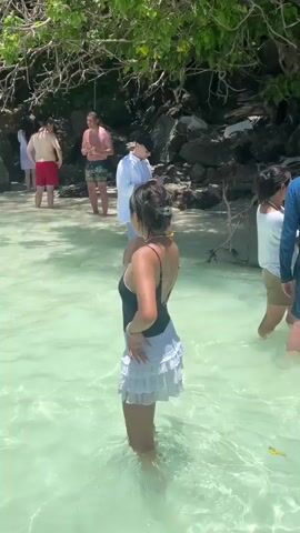 A group of people are gathered on a sandy beach with clear turquoise water. In the foreground, a young woman wearing a black swimsuit and a white skirt stands in the shallow water, facing away from the camera initially, then turns to smile. In the background, several other individuals are visible. One man in swim trunks holds up a phone, seemingly taking a photo. Another person in a white shirt also holds a phone. The environment appears to be tropical, with lush green foliage and rocky outcrops bordering the beach. The water is calm and reflects the sunlight.