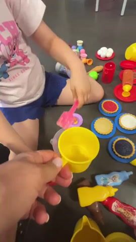 The video shows a close-up view of several children's toys scattered on a dark-colored floor. In the foreground, a person's hands are holding a yellow plastic cup. In the background, two children are partially visible, playing with the toys. One child, wearing a pink t-shirt and blue shorts, is reaching for a pink plastic utensil. The other child, mostly obscured, appears to be sitting and playing. The toys include miniature food items like pies and fruit, kitchen utensils, toy bottles, and a toy scale with fruit on it. The overall environment appears to be indoors, possibly a playroom or living area.