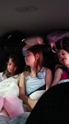 The video begins inside a vehicle where two young girls are seated in the back, surrounded by luggage. One girl smiles at the camera, while the other looks away. The camera then cuts to a woman with pink hair standing next to an open car door, adjusting her clothing. The perspective shifts to the driver's seat, showing the dashboard and a truck in the distance. Finally, the video shows a rearview mirror reflecting a colorful sunset and a truck driving on a road.