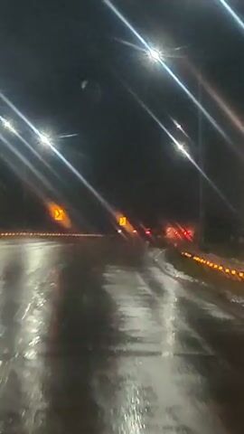 The video shows a first-person perspective of driving on a wet road at night. Streetlights with a bright glare illuminate the scene, casting reflections on the wet asphalt. Yellow road signs are visible on the left side of the road. In the distance, the taillights of other vehicles can be seen, indicating traffic. The overall impression is one of driving in rainy or wet conditions at night on a highway or a similar type of road.