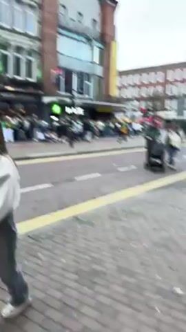 The video shows a crowd of people, many wearing green, gathered outside a bar called "White's Tavern" on a city street. People are walking on the sidewalks and across the street. A couple pushes a stroller along the sidewalk. The weather appears overcast. Buildings of varying heights and architectural styles line the street, and traffic is visible. A Mace convenience store is located near the bar.
