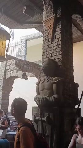 The video shows a crowd of people queuing outdoors in what appears to be a medieval-themed attraction or historical site. The architecture features stone walls, arched structures, and wooden beams, reminiscent of a castle or old town. A large, ornate lantern hangs from an overhead wooden beam, casting a warm glow. To the right, a stone statue of a knight in armor stands guard. In the background, a staircase leads up to a building with a red-tiled roof and wooden shutters. The people in the foreground are looking in various directions, some are talking, and others are simply waiting. The overall atmosphere suggests a tourist destination.
