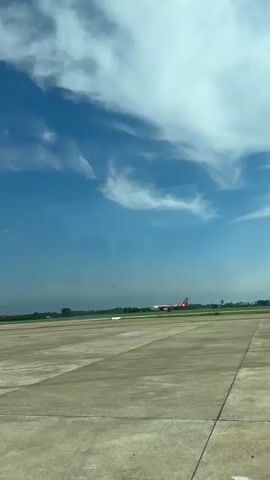 The video shows an Air Asia airplane with a red tail and a colorful livery on its side, taxiing on an airport tarmac. The airplane is positioned in the center of the frame, moving from left to right. The tarmac is made of concrete, and there is green grass on either side. In the background, there are trees and some buildings. The sky is blue with scattered white clouds. The video is shot from a slightly elevated perspective, likely from another aircraft or a building.