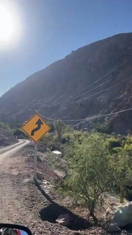 The video begins with a view of a winding dirt road in a mountainous desert landscape under a bright sun. A yellow diamond-shaped road sign with a black arrow indicating a sharp curve to the right is prominently displayed on a metal post in the foreground. To the left of the road, the terrain slopes upwards, revealing rocky mountainsides and sparse vegetation. Power lines crisscross the frame, supported by utility poles. On the right side of the road, the landscape descends into a valley with denser greenery, suggesting a water source. A small, primitive structure or shelter is partially visible amidst the vegetation. In the bottom left corner of the frame, a portion of a vehicle's side mirror reflects a blurred red object, possibly part of the vehicle's interior or a passenger. The camera appears to be mounted on or within this vehicle, suggesting a point-of-view shot. The overall impression is of travel through a remote, arid, yet surprisingly verdant canyon.
