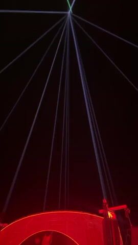 The video shows a stage setup at night with a dark background. Several blue laser beams emanate from a central point high in the sky, fanning out and downwards. Below, a large, modern stage structure is illuminated with bright red and orange lights. The stage features a prominent archway with reflective surfaces and red neon-like lighting. Inside the archway, there are several people standing on a platform. Speakers are mounted on the sides of the stage structure. The bottom of the frame shows a crowd of people gathered in front of the stage. The overall impression is a concert or performance setting with elaborate lighting and stage design.