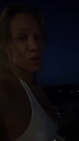 A woman with blonde hair is in a video, looking at the camera and pointing her finger towards the moon in the night sky. The background is dark with some faint lights visible in the distance. She is wearing a white tank top.