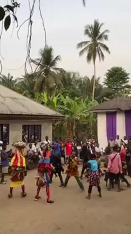 The video shows a group of people dancing in a dusty area in what appears to be a village. There are several people wearing colorful costumes, and a crowd of people watching. In the background, there are small buildings with corrugated metal roofs, and tropical trees including palm trees. Some people are standing and clapping. There is some kind of percussive music playing.