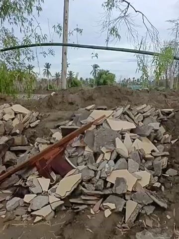 A pile of debris consisting of broken concrete, bricks, and ceramic tiles sits on the ground. Behind the debris, a tall utility pole stands with some bamboo trees to its left. In the background, there are more trees and what appears to be a rural landscape with some structures. The sky is overcast.