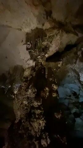 The video presents a close-up view of a geological formation inside a cave. The focus is on a column-like structure adorned with stalactites and stalagmites. The textures are rough and porous, with varying shades of brown, beige, and hints of white. Light sources illuminate sections of the formation, creating dramatic shadows that highlight the intricate details. The surrounding environment is dark, and the only other discernible features are parts of the cave ceiling and the floor, which are also rocky and textured. There is audio of speech, but no people are visible in the video.