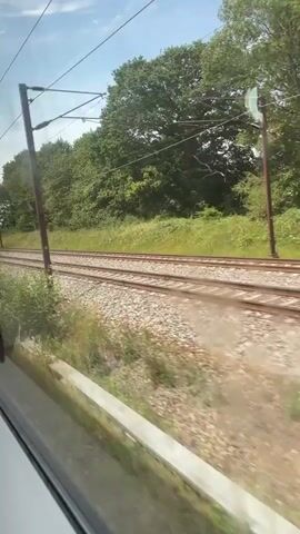 The video shows a view from inside a moving train looking out the window. The train tracks run parallel to the direction of travel, with gravel and vegetation on either side. Beyond the tracks, there is a line of trees and a blue sky with some clouds. Overhead, power lines and supporting poles are visible. The scene appears to be a rural or suburban landscape on a sunny day.
