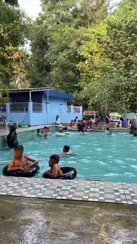 A group of people are enjoying a day at an outdoor swimming pool surrounded by lush green trees and a small blue building. Several children are playing in the water, some with inner tubes, while others are swimming and splashing. Adults are also present, supervising and relaxing by the poolside. The atmosphere appears to be cheerful and lively, indicating a recreational outing.