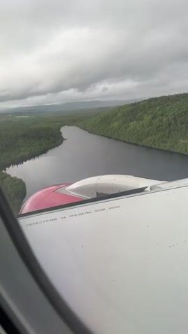 This video shows a view from an airplane window, looking out at a vast, green, forested landscape with a body of water winding through it. The sky is overcast with dark clouds. Part of an airplane wing and engine cowling are visible in the foreground, with Russian text printed on the wing. The landscape appears to be undeveloped and natural.