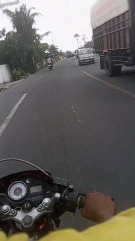 The video shows a first-person perspective of someone riding a motorcycle on a road. The rider's hand is visible on the handlebars, wearing a yellow sleeve. The motorcycle's speedometer is visible. The road is asphalt and appears to be in a populated area, with buildings and shops lining the right side of the road. Several cars and other motorcycles are also on the road, moving in the same direction. The sky is overcast.