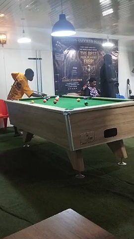 A man in an orange shirt is playing pool. He is standing on the left side of the pool table and is about to strike a ball with his cue. There are several balls on the table, including a white cue ball and several colored balls. Two chairs are visible behind the pool table, one red and one blue. Another person is seated in the foreground, facing away from the camera, with their legs crossed. A large fan is positioned to the left of the frame. The room is illuminated by overhead lights and a light fixture on the wall. A poster is visible on the back wall. The floor is covered with green carpet.
