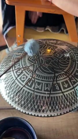 A close-up, eye-level shot shows a person's hand holding a malleted drumstick and striking a steel tongue drum. The drum is circular and metallic with an intricate, etched black pattern of geometric shapes and symbols. The malleted drumstick has a light blue, fluffy tip. The drum is resting on a wooden surface, with a dark blue bowl and a spoon visible in the foreground. In the background, a person's legs are visible, and they appear to be seated on a wooden stool with an orange frame. The overall environment suggests an indoor setting, possibly a home or a studio. The video captures the action of playing the musical instrument.